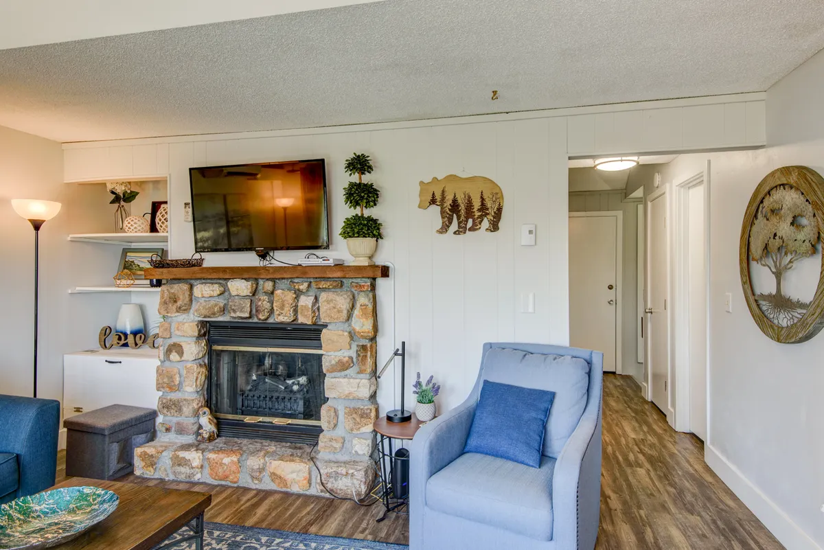 A cozy living room featuring a prominent stone electric fireplace, a plush blue armchair, decorative bear-themed artwork, and a touch of greenery. The space is arranged for relaxation and gathering, with a warm ambiance ideal for winter evenings.