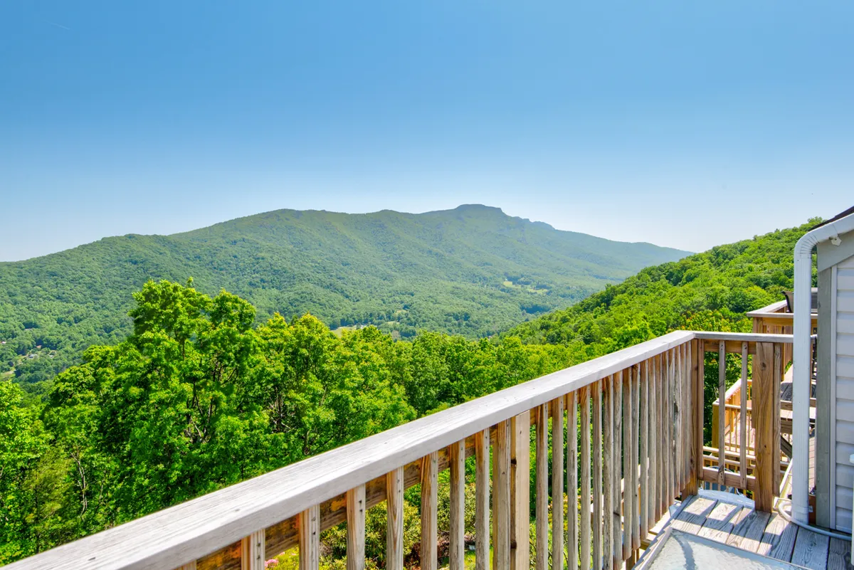 A serene view from a wooden balcony overlooking mountains and verdant hills, framed by lush greenery. The perspective invites viewers to imagine sipping coffee or watching sunrise before heading out for the day's adventures.