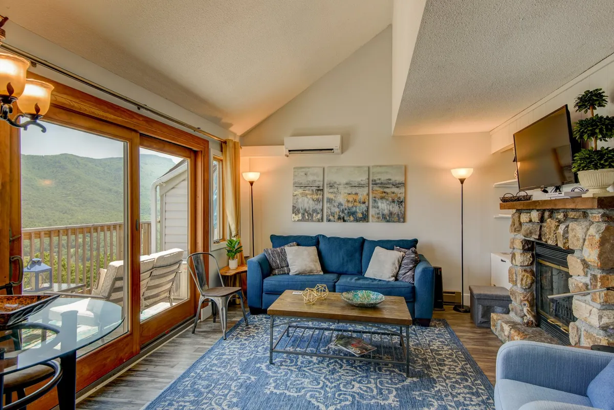 An inviting living room with a large, soft sofa and a modern fireplace, framed by bright windows offering glimpses of the mountain landscape outside. The cozy coffee table and artful decor create an atmosphere ideal for relaxation, socializing, or watching TV with loved ones.
