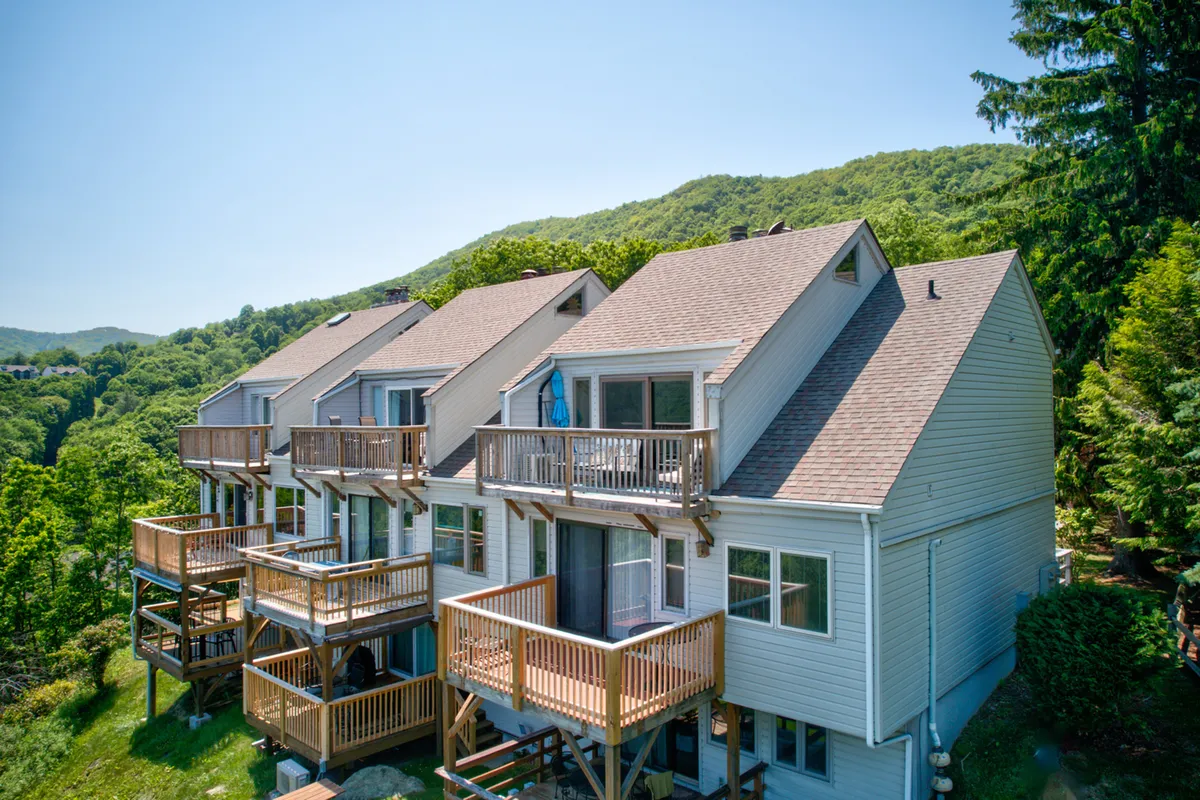 The image features the condo’s wooden balconies overlooking a stunning mountain landscape, with Grandfather Mountain visible in the distance. The grounds are neatly landscaped, and the elevated perch offers panoramic views perfect for relaxing or gathering at sunset. The overall mood is tranquil and inviting, showcasing the property’s elevation and natural beauty.