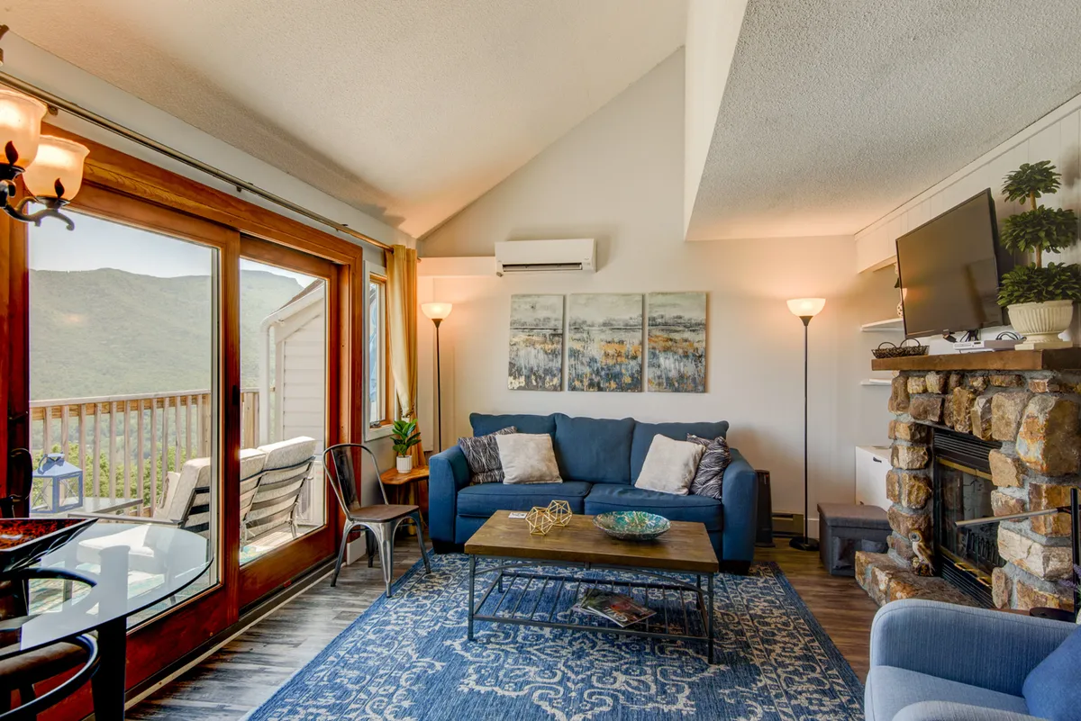 A cozy living room featuring a comfortable blue sofa positioned to enjoy panoramic mountain views through expansive windows. A striking stone fireplace anchors the space, while tasteful artwork and a wooden coffee table add warmth and personality. Natural light fills the room, showcasing the inviting ambiance and the ever-present beauty of Grandfather Mountain.