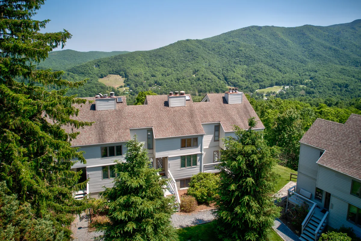 A peaceful exterior shot showing the building set against a beautiful mountain backdrop, surrounded by lush greenery. The environment suggests tranquility and access to nature, ideal for gentle hikes and outdoor exploration.