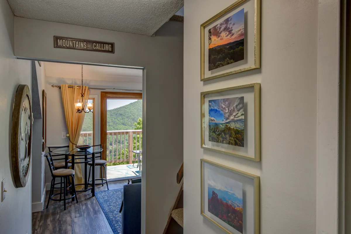 A stylish dining area inside, set near a large doorway that frames breathtaking mountain views and a private balcony. The dining table offers a scenic spot for meals or morning coffee, complemented by framed photographs and soft natural light.