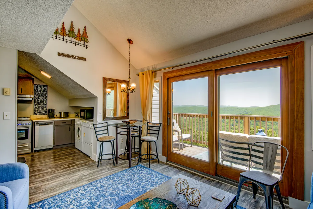 An open-plan living and dining area with large sliding glass doors leading to the balcony, a blue area rug, kitchenette, dining bar with stools, and stylish wall art. The setup encourages both relaxation and socializing, with abundant natural light and an easy flow to the outdoors.