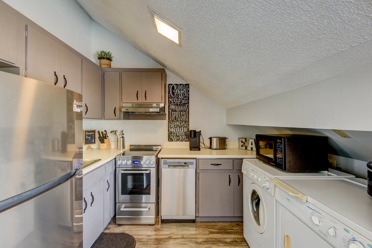 A practical interior featuring stainless steel appliances, dark cabinetry, decorative wall art, and a visible laundry machine tucked into the kitchen area. The setting is both inviting and functional, perfect for handling laundry and meal prep with ease.