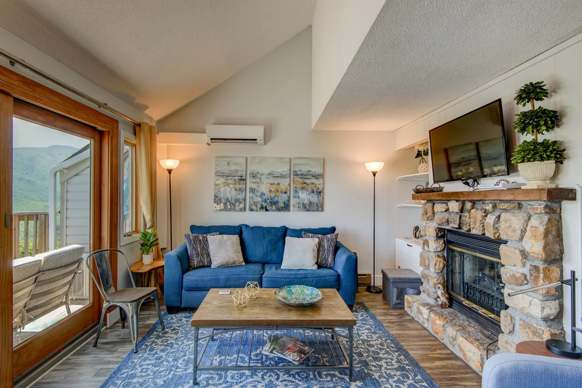 A warm, welcoming living area with a blue sofa, stone fireplace, coffee table, wall art, and large windows letting in natural light. The arrangement is perfect for relaxing with a book, enjoying friendly conversations, or sipping a warm drink by the fire.