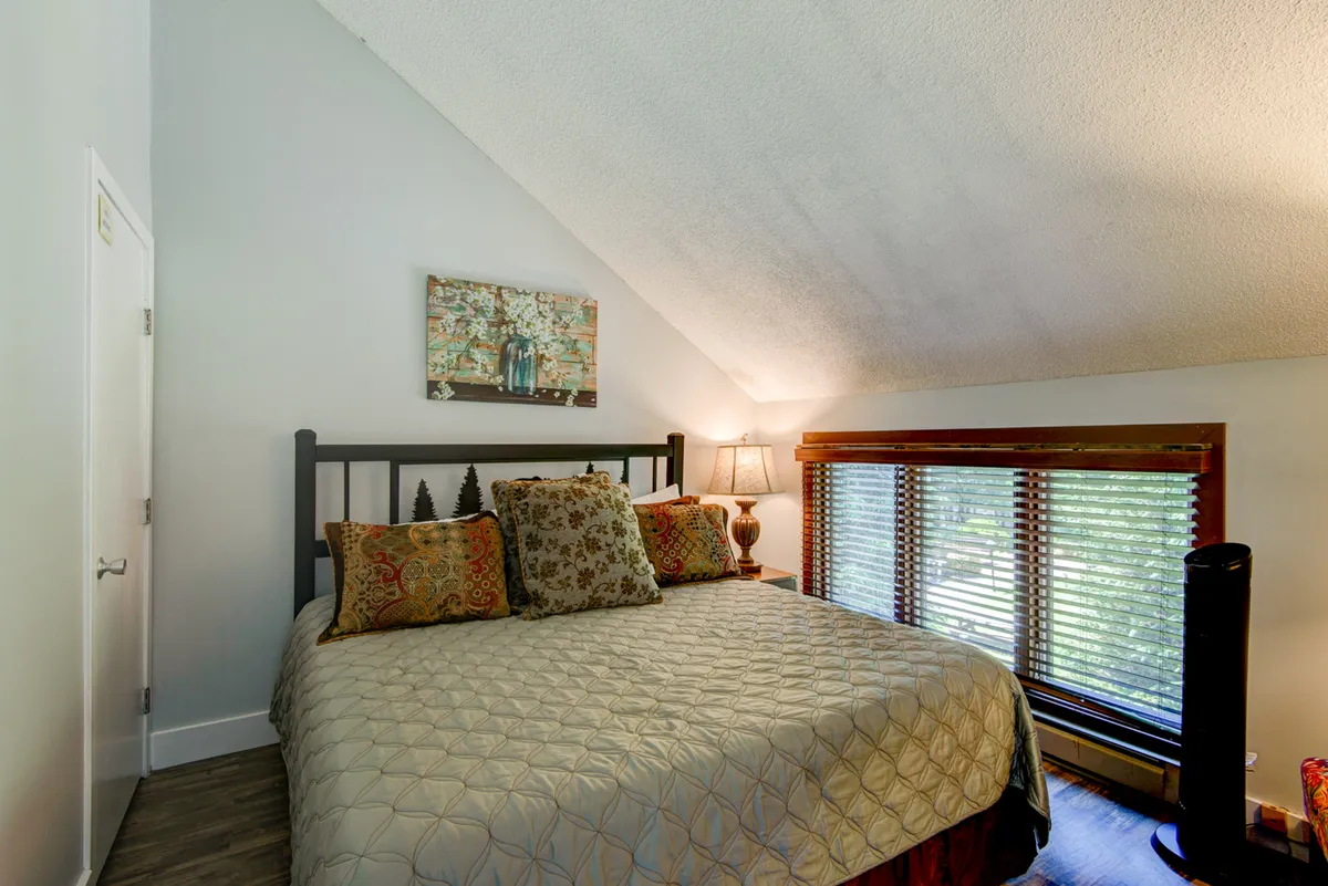 A cozy bedroom with a large bed made up with decorative pillows, a piece of artwork hanging above the headboard, and a soft-glow table lamp beside the bed. The overall lighting and decor evoke comfort and privacy, making it ideal for relaxation after a long day.