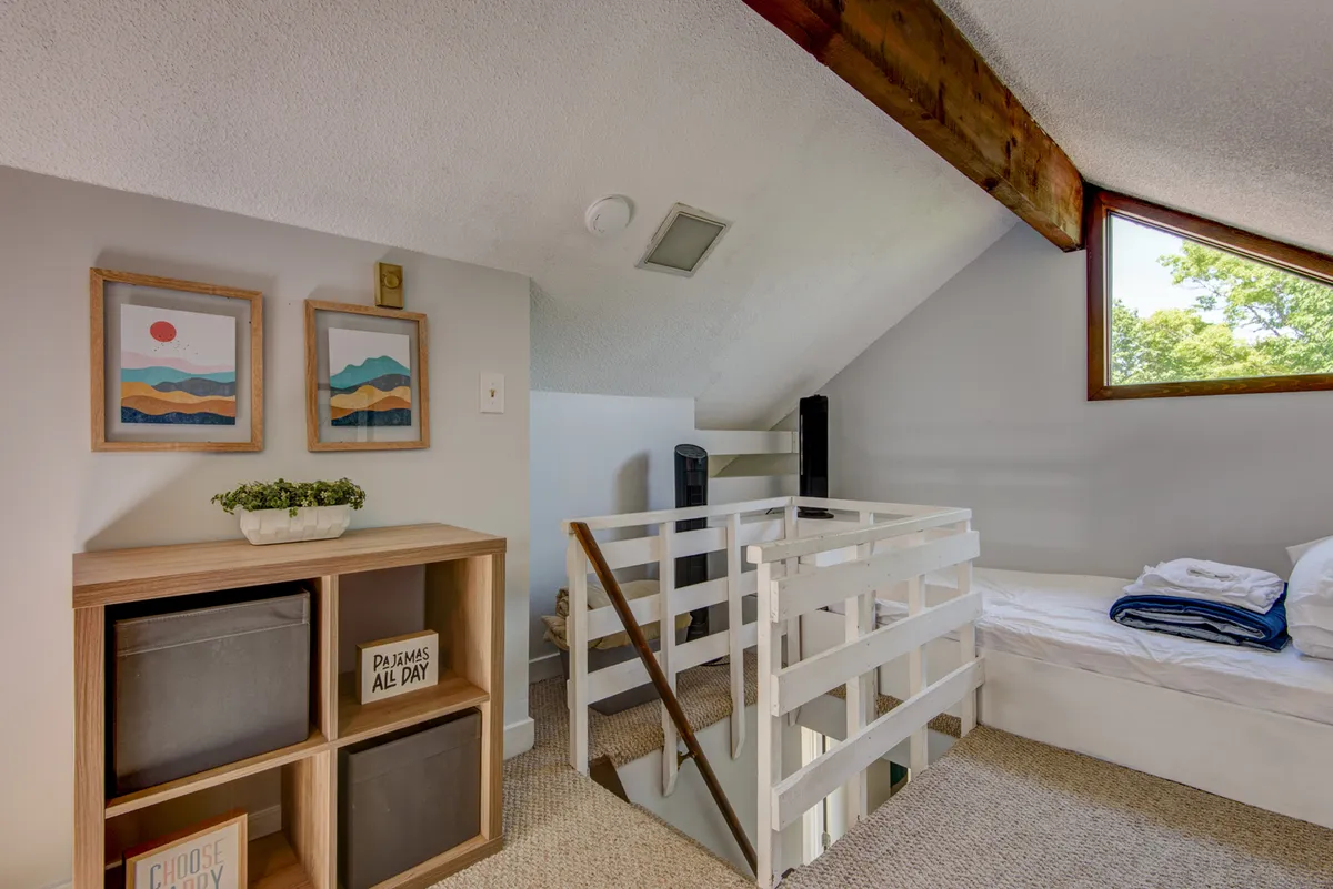 An intimate loft sleeping nook featuring a bed dressed in simple linens, a rustic wooden beam, shelves for storage, and a window offering natural light. The nook’s compactness and the ladder-accessible location create a sense of adventure and coziness, ideal for kids or the young at heart.